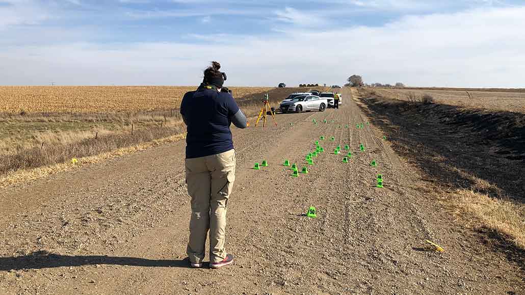 a photo showing Samantha Hayek facing away down a dirt road speckled with green evidence flags. She is taking photos of the scene.