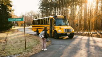 the early morning sun peeks through the trees as a middle school girl stands on the curb waiting for an approaching school bus