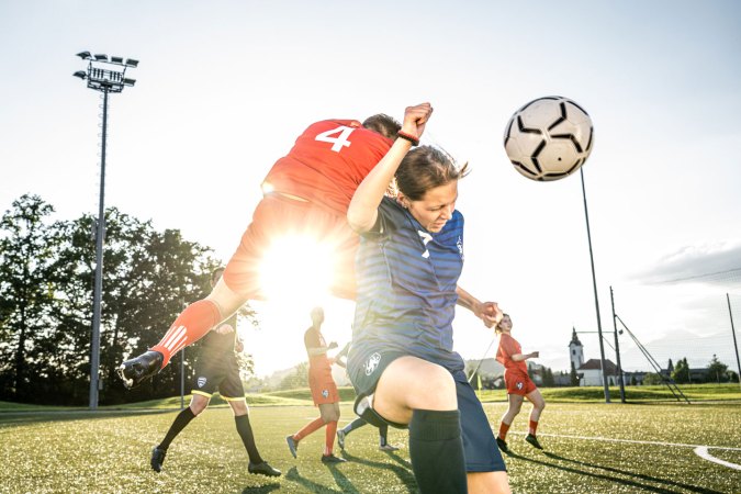one teen soccer player on her knee throws her arm up to block another player about to collide with her back