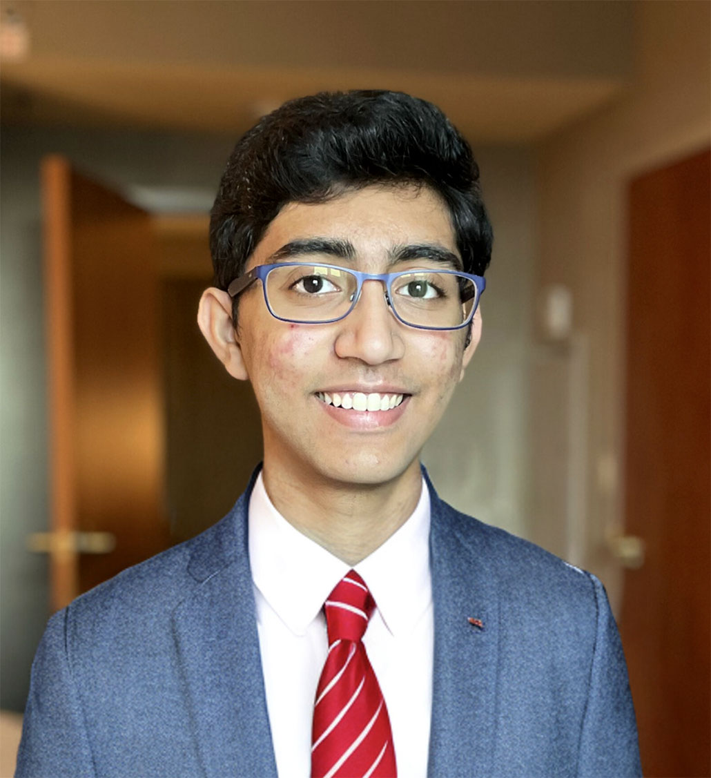 a photo of Saathvik Kannan, a young man with dark hair, glasses, brown skin and a big smile. He is wearing a grey suit and a red tie.