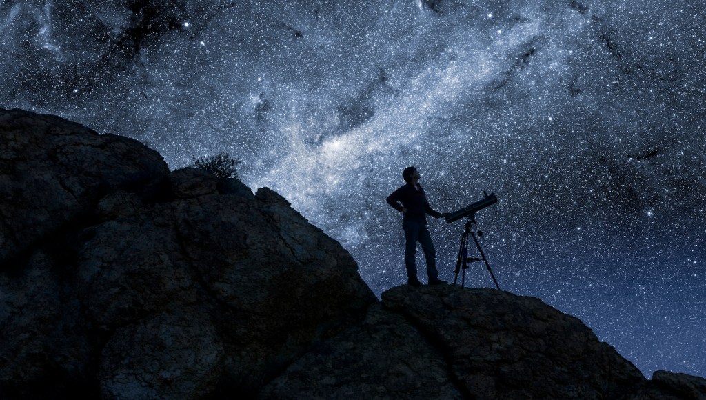 A person with a telescope standing on a dark hillside, silhouetted against a starry night sky.
