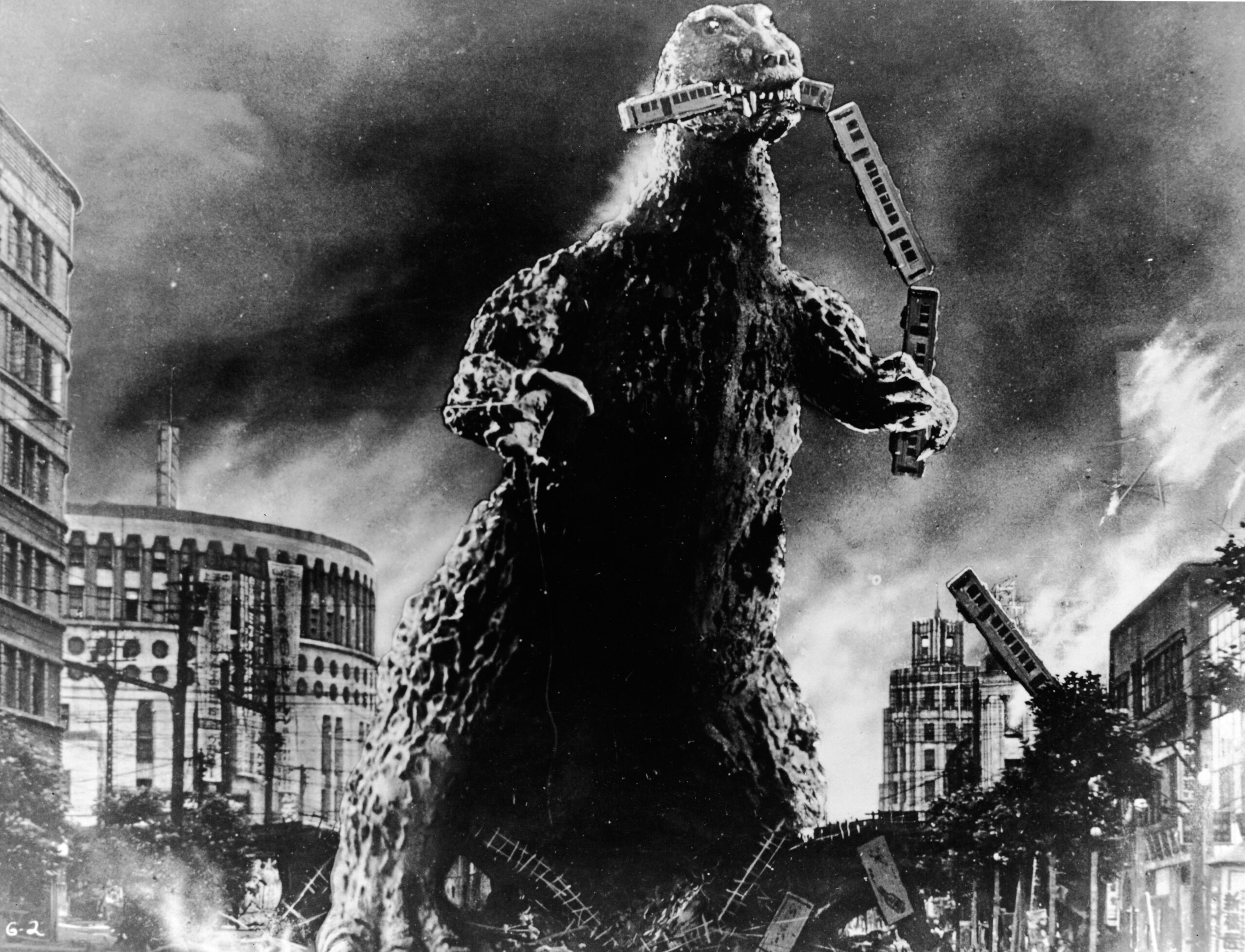 Godzilla holds a commuter train in his mouth. A ruined city surrounds him.