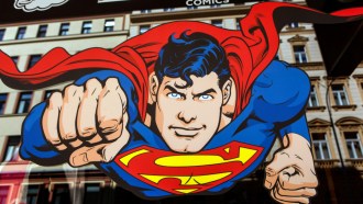 An illustrated Superman flies head on toward the viewer. He is a white man with black hair and blue eyes. His costume sports a yellow diamond with a red S in the middle. His red cape trails behind him. A tan, multistory building is in the background