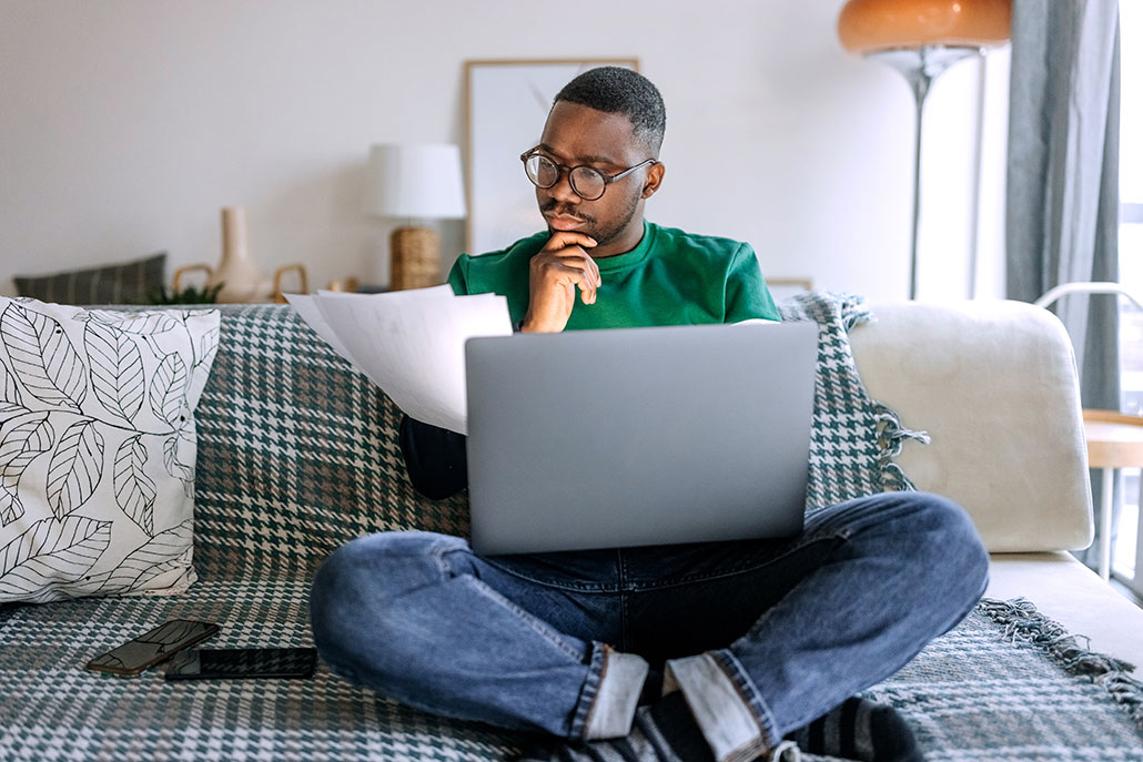a young black man with short hair and glasses is sitting on a sofa with a laptop. He's holding a stack of papers and reading them.