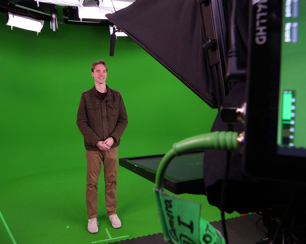 a photo of a young white man standing in a studio with green walls and floor, being filmed