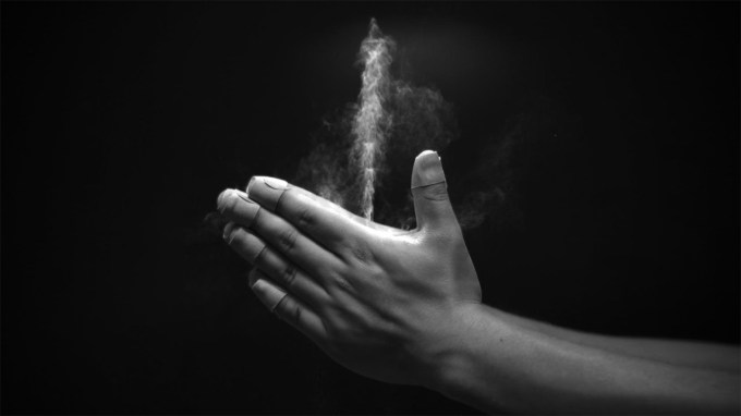 A grayscale images shows two hands clapping together, with a white jet of baby powder streaming upward from a space between the thumbs and forefingers.