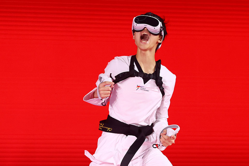 a photo of an Asian teen wearing a taekwondo outfit and a VR headset and gear yelling in victory in front of a red background. His eyes are covered by the VR set.