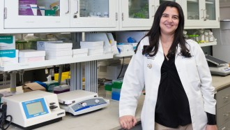 Hearing researcher A. Catalina Vélez-Ortega stands next to a counter with scientific machines. She is wearing a white lab coat and has black hair and fair skin. White shelves line the walls behind her.