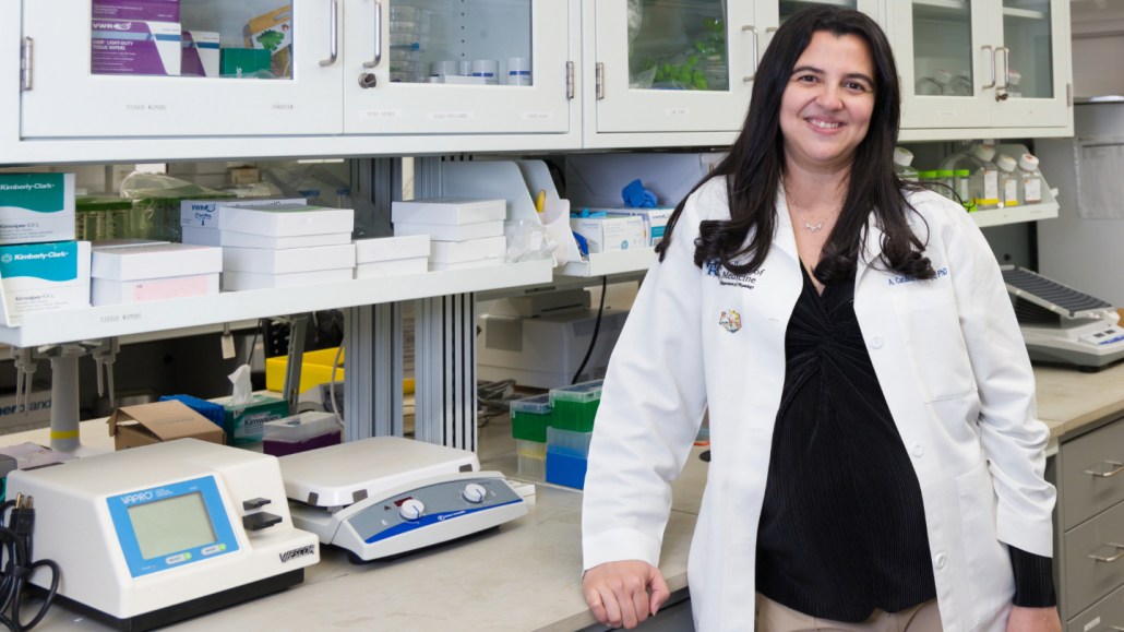 Hearing researcher A. Catalina Vélez-Ortega stands next to a counter with scientific machines. She is wearing a white lab coat and has black hair and fair skin. White shelves line the walls behind her.
