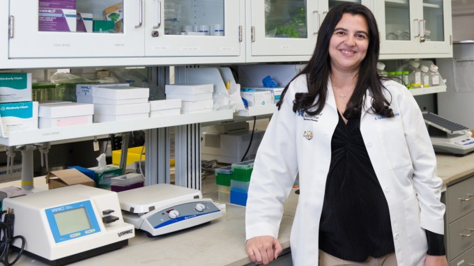 Hearing researcher A. Catalina Vélez-Ortega stands next to a counter with scientific machines. She is wearing a white lab coat and has black hair and fair skin. White shelves line the walls behind her.
