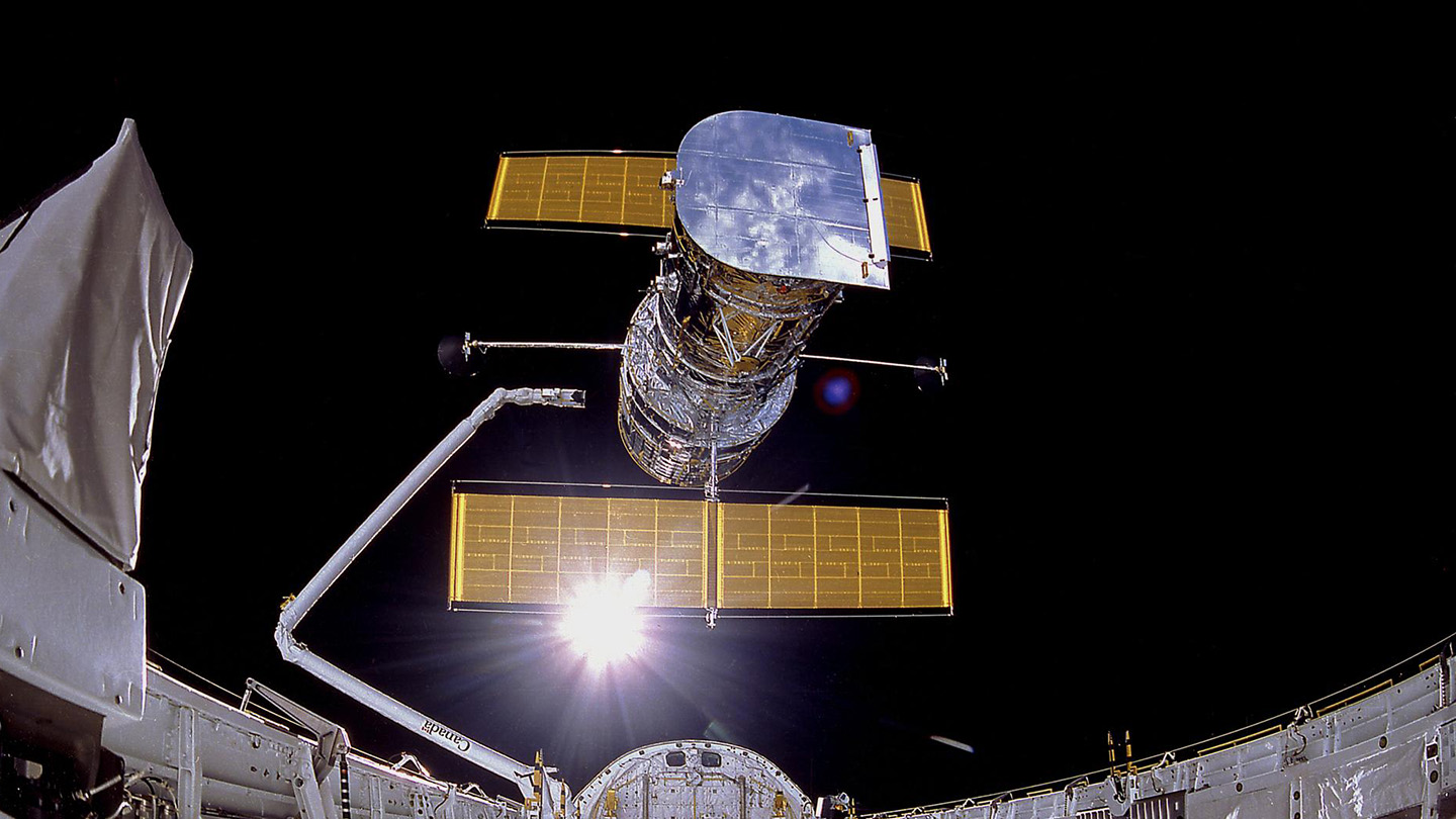 The Hubble Space Telescope being deployed from a space shuttle into orbit, with solar panels extended against the black backdrop of space and the sun shining brightly behind the observatory.