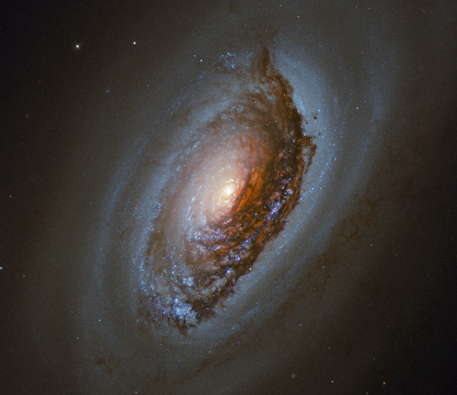 A spiral of light dotted with stars with a dark, reddish brown splotch stretching across much of the spiral. The center of the spiral glows with yellowish light.