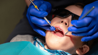 A child in sunglasses has dental tools inserted in mouth by practitioner wearing blue gloves.