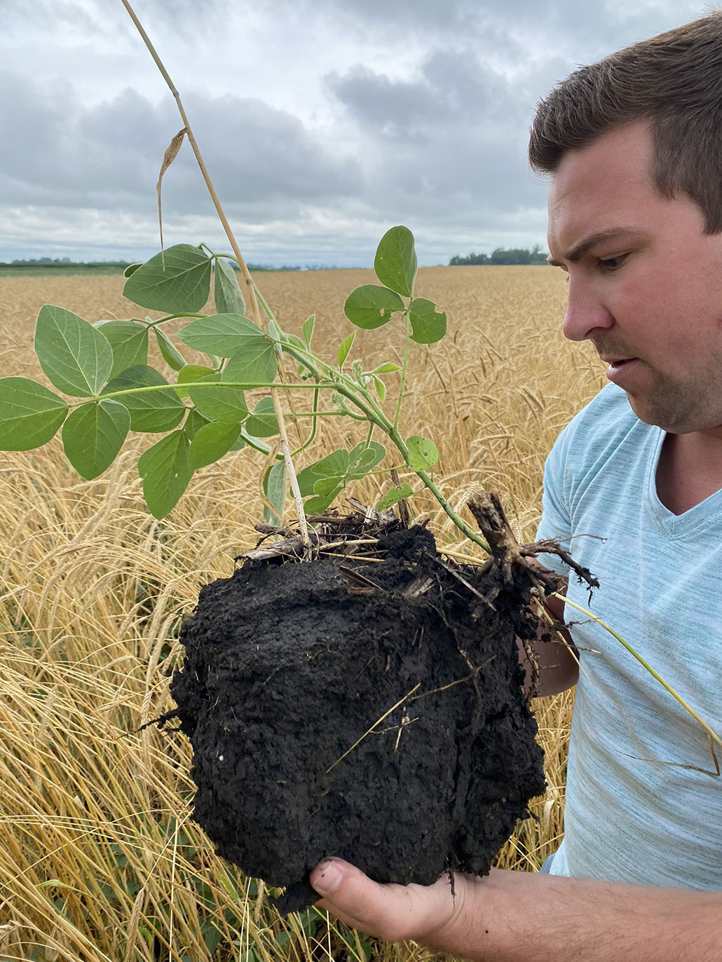 Mitchell Hora, a white man with light colored hair, is seen standing in a field holding a large ball of health soil with a plant emerging from the top of it