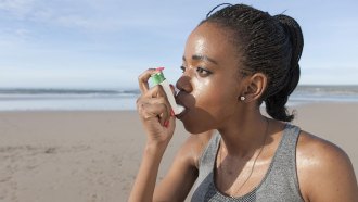 a woman is using an inhaler at a beach, she has dark skin and dark hair in microbraids and a ponytail. She seems to have been excercising.