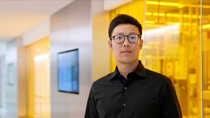 Molecular engineer Pengju Li stands in a hallway, in front of yellow tinted glass. He has black hair and brown eyes. He is wearing black-framed glasses and a black dress shirt.
