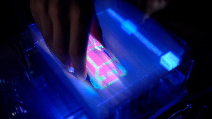 a hand reaches down into an illuminated screen displaying a virtual 3-D graphic of a car