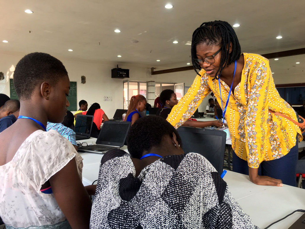 Tabiri leans over a desk in a classroom helping students. She's wearing glasses, chin length braids and a yellow shirt. 