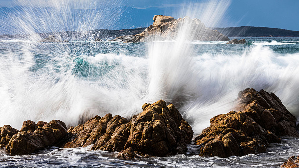 a photo of ocean water hitting rocks at a shoreline and spraying water vertically into the air in a white sheet