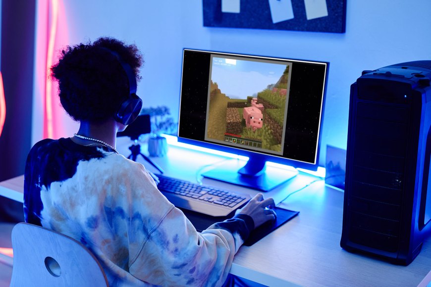 a teen sits in front of a computer looking at a screen with a video game scene AI-generated by Oasis, featuring a Minecraft-like pig. The teen is wearing a black, white and cream tie-dyed hoodie. They have dark brown curly hair and dark skin. Although the room is dark, there are rgb lights behind the monitor and along the wall illuminating the gaming space.