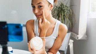 a young woman holding a skincare product and applying a cream to her face while sitting in front of a smartphone on a tripod