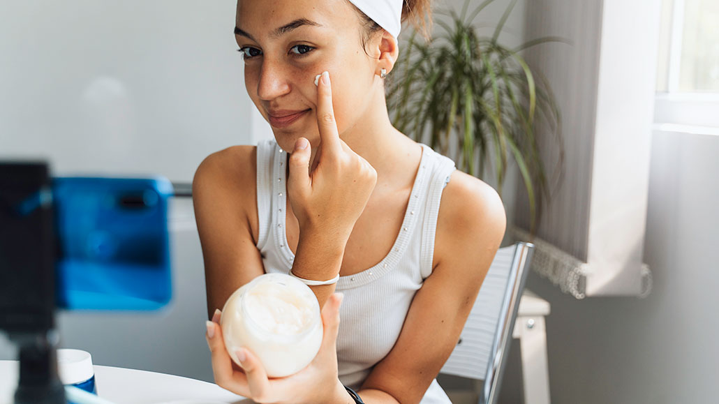 a young woman holding a skincare product and applying a cream to her face while sitting in front of a smartphone on a tripod