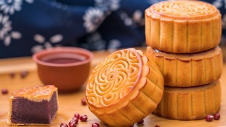 A stack of brown mooncakes sit on a wooden table. Red beans are scattered nearby.