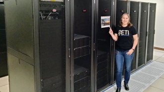 Chenoa Tremblay stands in front of a row of supercomputer servers. She is a white woman with brown, shoulder-length hair. She is wearing blue jeans and a black T-shirt with "SETI" spelled out on the front.