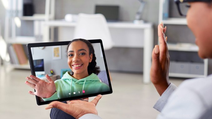 a person seen in profile is wearing black rimmed glasses and waving at their friend on some type of screen technology. The friend seems to pop out of the screen like a hologram.