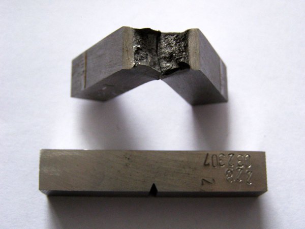 a small piece of metal with a notch in it is at the bottom, at the top the same metal sample after a charpy test (the metal has broken in half)