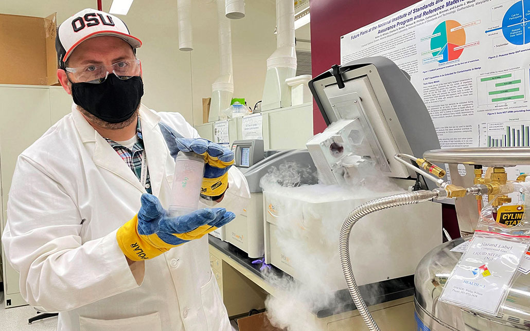 a man in a white lab coat wearing thick gloves is about to place a sample into a cryogrinder