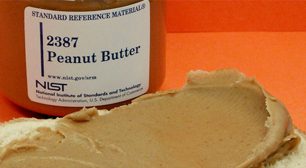 a jar of peanut butter labeled 'Standard Reference Material, 2387 Peanut Butter, NIST' against an orange backdrop. A smear of peanut butter is spread on bread in front of the jar.