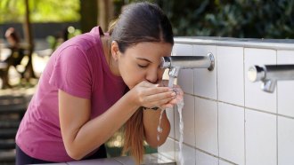 a woman cups her hands to drink water from a water fountain in a park