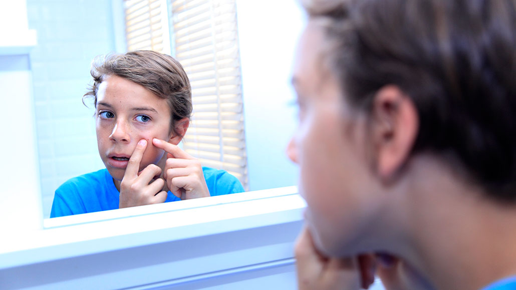 a boy with light skin and light brown hair examines a red blemish on his face in the mirror